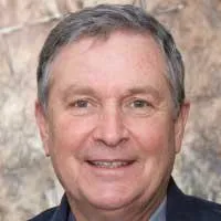 A middle-aged man with short gray hair, wearing a navy blazer, stands against an abstract textured background.